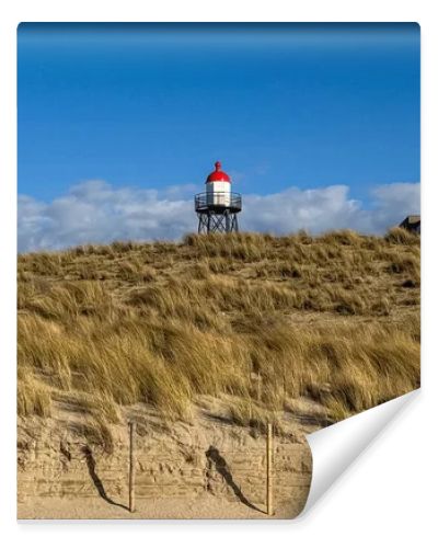 Scenic Dutch dune landscape featuring sandy hills, dry grass and a red and white lighthouse beneath bright clouds and blue sky. Peaceful coastal nature scene suitable for travel, tourism and environmental concepts.