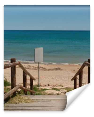Wooden beach access path leading to a sandy Mediterranean shoreline with calm blue sea and blank white signboard for copy space or custom design. Sunny coastal scene with natural seaside atmosphere.