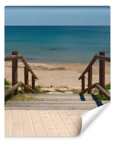 Wooden boardwalk with railings leading to a quiet sandy beach and turquoise Mediterranean sea. Sunny coastal scene with clear blue sky and peaceful vacation atmosphere.
