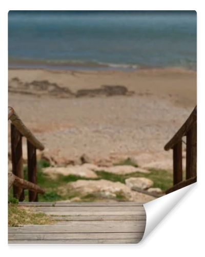 Wooden boardwalk with railings leading to a quiet sandy beach and turquoise Mediterranean sea. Sunny coastal scene with clear blue sky and peaceful vacation atmosphere.