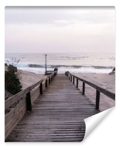 A serene beach scene in Portugal. A wooden boardwalk through sand dunes to the ocean, where waves lap the shore under a cloudy sky. Sparse vegetation and evening light create a tranquil atmosphere.