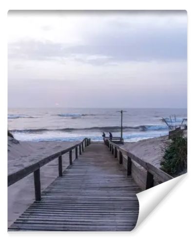 A serene beach scene in Portugal. A wooden boardwalk through sand dunes to the ocean, where waves lap the shore under a cloudy sky. Sparse vegetation and evening light create a tranquil atmosphere.