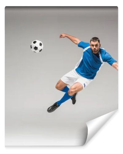 Sportsman looking at camera while jumping near football on grey background