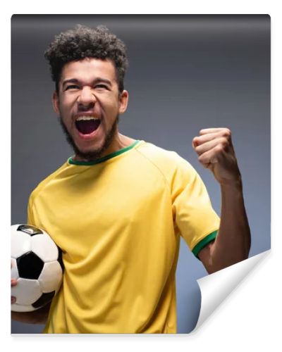 excited male african american football fan in yellow t-shirt holding ball on grey