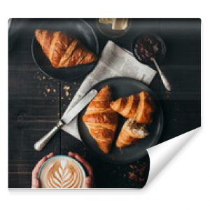 Hands holding latte beside croissants on black table from above.