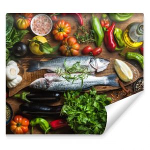 Raw uncooked seabass fish with vegetables, grains, herbs and spices on chopping board over rustic wooden background