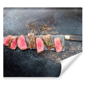 Traditional barbecue aged venison backstrap roast sliced with herbs as top view on a large knife with rustic background with copy space