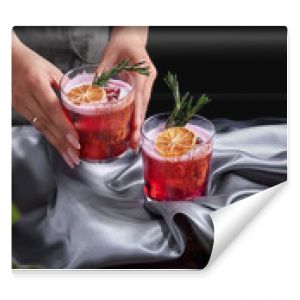 Red drink cocktail of rum and mezcal with lime and cherry juice garnished with dried fruit and green rosemary placed by two female hands