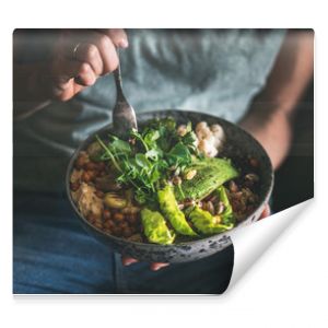 Healthy dinner or lunch. Woman in t-shirt and jeans eating vegan superbowl or Buddha bowl with hummus, vegetable, salad, beans, couscous and avocado