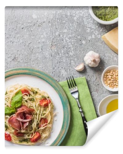 top view of cooked Pappardelle with tomatoes, basil and prosciutto near ingredients, red wine and cutlery on grey surface