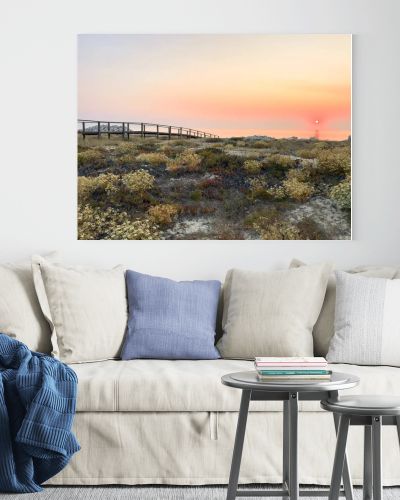 A wooden boardwalk stretches across wild coastal dunes blooming with flora towards a radiant golden sunset over the calm ocean horizon at Furadouro beach in Ovar Portugal creating a serene scene