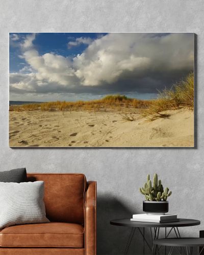 Sandy dunes dotted with grass stretch toward a distant ocean horizon beneath dramatic storm clouds, evoking solitude, nature, and changing weather on a quiet coastal landscape at low angle light.
