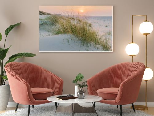 Golden hour paints the sky above the serene beach and dunes. Tall grasses sway gently, casting soft shadows on the cool, blue sand as the sun dips below the horizon.