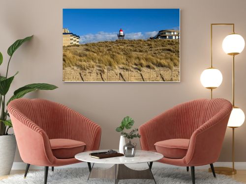 Scenic Dutch dune landscape featuring sandy hills, dry grass and a red and white lighthouse beneath bright clouds and blue sky. Peaceful coastal nature scene suitable for travel, tourism and environmental concepts.