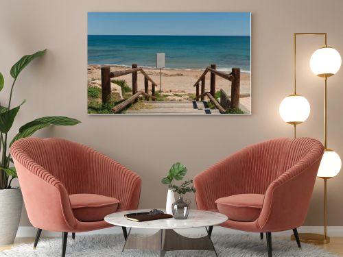 Wooden beach access path leading to a sandy Mediterranean shoreline with calm blue sea and blank white signboard for copy space or custom design. Sunny coastal scene with natural seaside atmosphere.