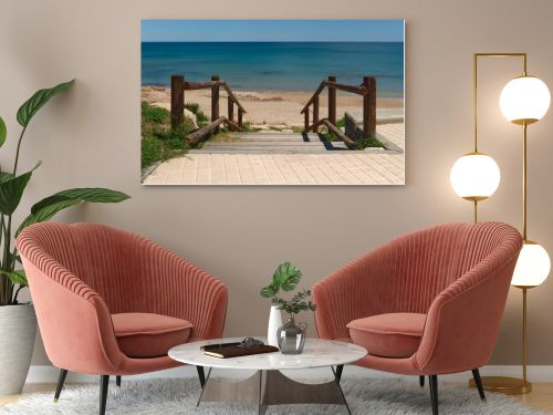 Wooden boardwalk with railings leading to a quiet sandy beach and turquoise Mediterranean sea. Sunny coastal scene with clear blue sky and peaceful vacation atmosphere.