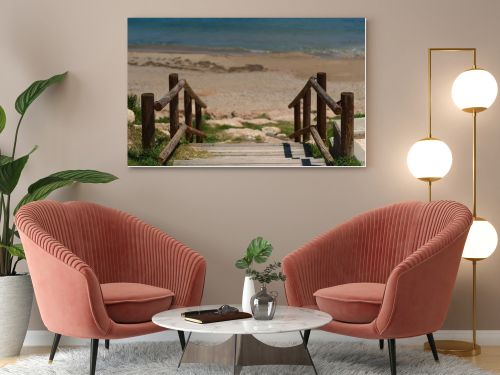 Wooden boardwalk with railings leading to a quiet sandy beach and turquoise Mediterranean sea. Sunny coastal scene with clear blue sky and peaceful vacation atmosphere.