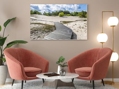 Wooden boardwalk leads over white coastal dunes in Latvia, framed by shrubs, pines and dramatic clouds.
