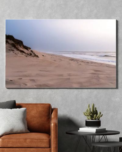 A serene, sandy beach with gentle waves against the shore under a soft, pastel sky. Dunes with sparse vegetation rise gently from the beach, creating a peaceful and untouched natural landscape.