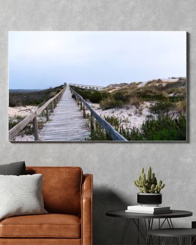 A serene beach scene in Portugal. A wooden boardwalk through sand dunes to the ocean, where waves lap the shore under a cloudy sky. Sparse vegetation and evening light create a tranquil atmosphere.
