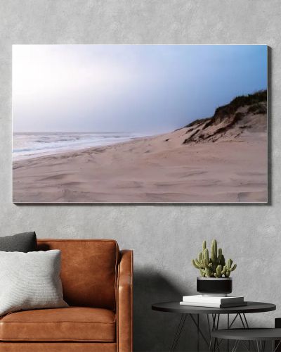 A serene, sandy beach with gentle waves against the shore under a soft, pastel sky. Dunes with sparse vegetation rise gently from the beach, creating a peaceful and untouched natural landscape.