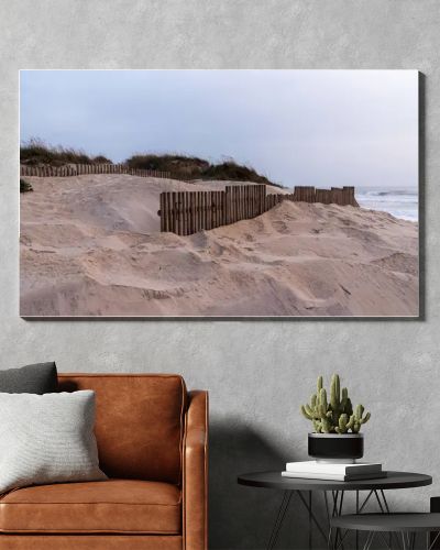 A coastal dune landscape in Nazare, Portugal, with sand dunes stabilized by wooden fences and sparse vegetation. The overcast sky and waves of the Ocean create a serene and natural seaside atmosphere