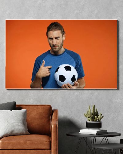 football fan in blue t-shirt holding soccer ball and pointing at himself on orange 