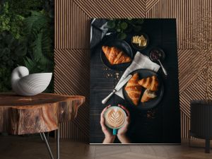 Hands holding latte beside croissants on black table from above.