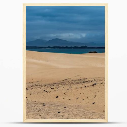 Spain, Canary Islands, Fuerteventura-February 5, 2026: Golden sand dunes stretch toward blue ocean and volcanic mountains beneath cloudy sky, forming dramatic coastal desert landscape.
