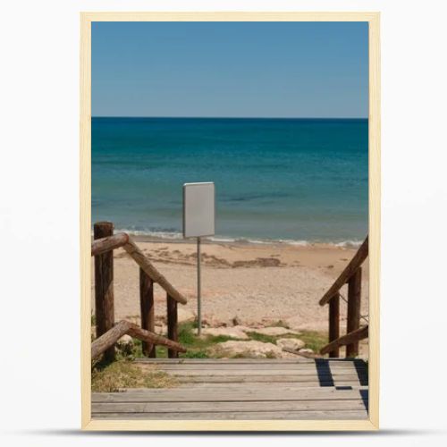 Wooden beach access path leading to a sandy Mediterranean shoreline with calm blue sea and blank white signboard for copy space or custom design. Sunny coastal scene with natural seaside atmosphere.