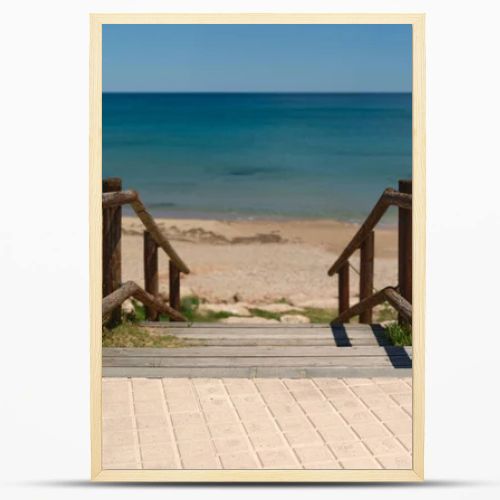 Wooden boardwalk with railings leading to a quiet sandy beach and turquoise Mediterranean sea. Sunny coastal scene with clear blue sky and peaceful vacation atmosphere.