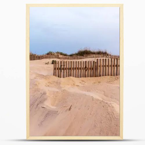 A sandy dune landscape with wooden fences lines the edge of a beach under an overcast sky. The scene captures the raw beauty of a coastal environment with a serene and untouched atmosphere.