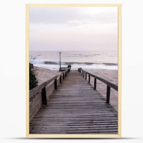 A serene beach scene in Portugal. A wooden boardwalk through sand dunes to the ocean, where waves lap the shore under a cloudy sky. Sparse vegetation and evening light create a tranquil atmosphere.