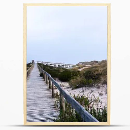 A serene beach scene in Portugal. A wooden boardwalk through sand dunes to the ocean, where waves lap the shore under a cloudy sky. Sparse vegetation and evening light create a tranquil atmosphere.