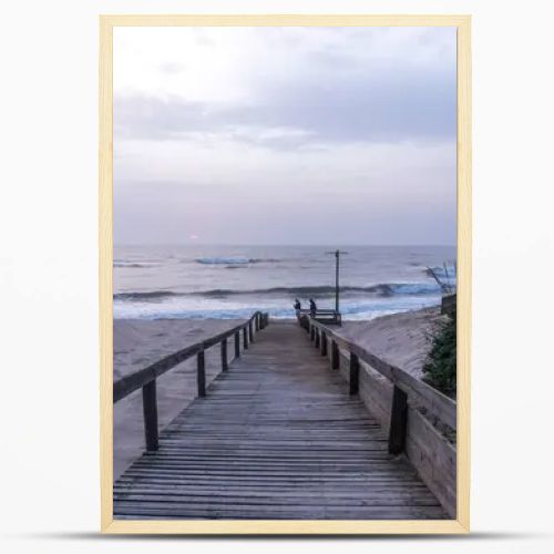 A serene beach scene in Portugal. A wooden boardwalk through sand dunes to the ocean, where waves lap the shore under a cloudy sky. Sparse vegetation and evening light create a tranquil atmosphere.