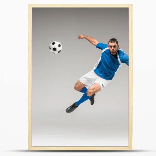 Sportsman looking at camera while jumping near football on grey background