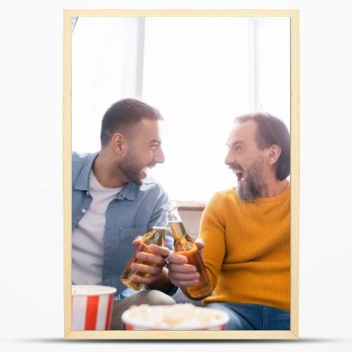 excited interracial dad and son clinking bottles of beer while watching football championship at home