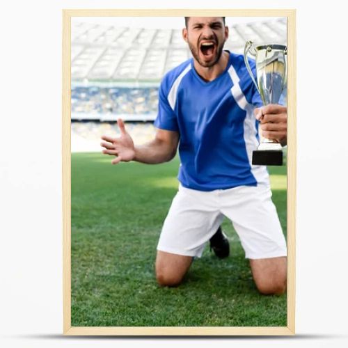 professional soccer player in blue and white uniform with sports cup standing on knees on football pitch and shouting at stadium