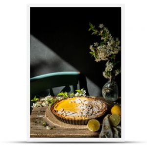 Freshly baked homemade lemon pie with floral crust decorations  in baking form, lemons and cherry blossom  on old wooden table on dark background. 