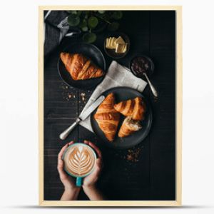 Hands holding latte beside croissants on black table from above.