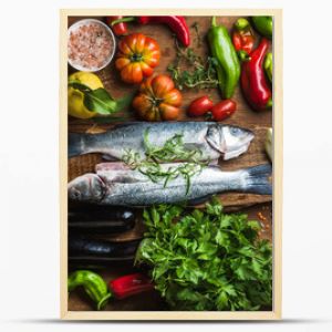 Raw uncooked seabass fish with vegetables, grains, herbs and spices on chopping board over rustic wooden background