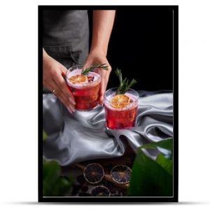 Red drink cocktail of rum and mezcal with lime and cherry juice garnished with dried fruit and green rosemary placed by two female hands