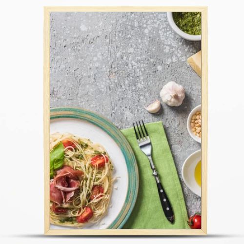 top view of cooked Pappardelle with tomatoes, basil and prosciutto near ingredients, red wine and cutlery on grey surface