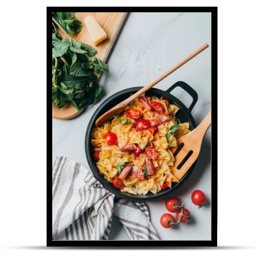 elevated view of pasta with jamon, cherry tomatoes, mint leaves covered by grated parmesan in pan with wooden spatula and spoon on marble table