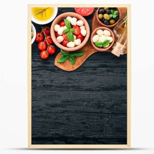 Ingredients for Italian caprese salad. Mozzarella cheese, cherry tomatoes, basil leaves, olives, oil, pepper. On a black wooden background. Free space for text.