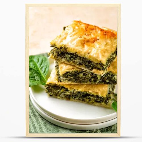 Stack of Spanakopita piece of pie, homemade Greek savory spinach pastry. It contains cheese feta, chopped spinach, green, egg, layered in phyllo or filo dought. Light beige background with green napkin.