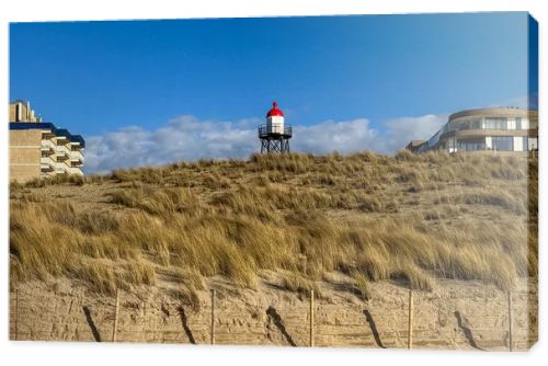 Scenic Dutch dune landscape featuring sandy hills, dry grass and a red and white lighthouse beneath bright clouds and blue sky. Peaceful coastal nature scene suitable for travel, tourism and environmental concepts.