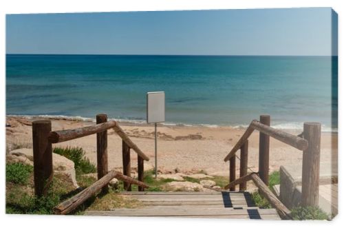 Wooden beach access path leading to a sandy Mediterranean shoreline with calm blue sea and blank white signboard for copy space or custom design. Sunny coastal scene with natural seaside atmosphere.