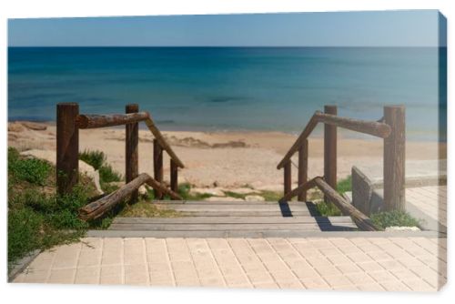 Wooden boardwalk with railings leading to a quiet sandy beach and turquoise Mediterranean sea. Sunny coastal scene with clear blue sky and peaceful vacation atmosphere.