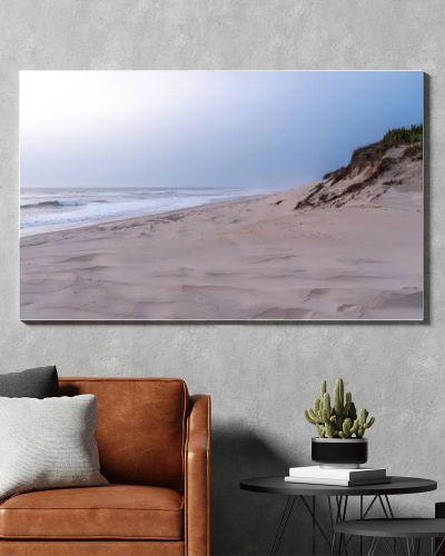 A serene, sandy beach with gentle waves against the shore under a soft, pastel sky. Dunes with sparse vegetation rise gently from the beach, creating a peaceful and untouched natural landscape.